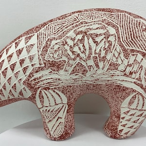 May include: A reddish-brown bear sculpture with white etched designs. The bear has a detailed carving of a bear on its back, and geometric patterns on its body. The sculpture is a decorative item.