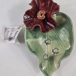 May include: A handcrafted ceramic leaf-shaped dish with a dark red flower accent. The dish is green with three turtle designs. The flower has a yellow center. The tag reads "Pottery by Kaivni".