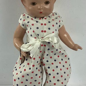 May include: Vintage doll with a painted face and auburn hair, wearing a white jumpsuit patterned with red and navy blue polka dots. The jumpsuit has a white satin bow at the waist. The doll is wearing white socks.