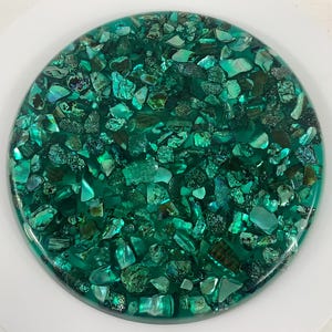 May include: A round, teal-colored decorative piece with embedded iridescent shell fragments. The shell pieces vary in size and shape, creating a textured surface. The piece has a glossy finish and is set against a white background.