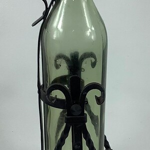 May include: A decorative green glass bottle with a cork stopper, held in a black wrought iron stand. The stand has a handle and ornate scrollwork. The bottle is approximately 12 inches tall.