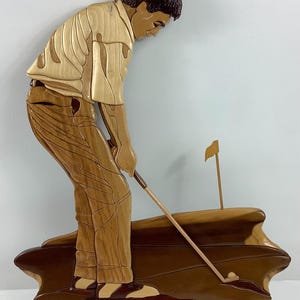 May include: Wooden wall art depicting a golfer in action, crafted from various wood tones. The figure is dressed in a light-colored shirt and brown pants, poised to swing. A golf ball and flag are also visible. The artwork is a unique piece of home decor.