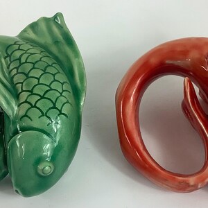May include: Two ceramic fish-shaped napkin rings. One is a glossy green, the other is a glossy red-orange. The fish have a curved body and a detailed tail fin. The fish scales are visible. These decorative rings are for table settings.