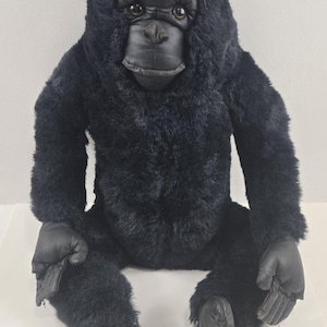 May include: A plush gorilla toy featuring thick, black faux fur covering its body, arms, and legs. The face, hands, and feet are made of a smooth, black material. The gorilla has dark, round eyes and a neutral facial expression, sitting upright.