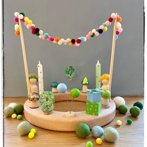 May include: A circular wooden birthday ring featuring a colorful pom-pom garland suspended between two wooden dowels. The ring is decorated with wooden peg dolls, a glittery number nine, two candles, miniature mushrooms, and small gift boxes.