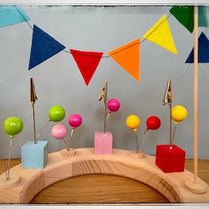 Può includere: Un arco in legno arcobaleno con bandierine di feltro colorate e piccoli palloncini colorati attaccati a delle clip. I palloncini sono attaccati a blocchi di legno di vari colori.