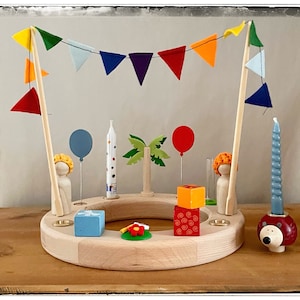 Op de afbeelding: Een houten verjaardagstaart met kleurrijke vlaggen van vilt, een blauwe ballon van vilt, een houten palmboom en een houten figuur met een gele hoed. Er zijn ook houten kaarsen, een blauwe doos van vilt en een rode doos van vilt.