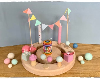May include: A circular wooden birthday ring featuring a central candle with rainbow stripes and the text HAPPY BIRTHDAY. The ring is decorated with two tall candles, a pastel bunting banner, a wooden pig, two mushrooms, and two gift boxes.
