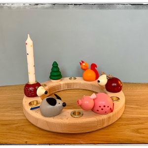May include: Wooden advent calendar with a white candle, a red squirrel, a pink pig, a grey mouse, a brown dog, and a green tree. The calendar has four candle holders.
