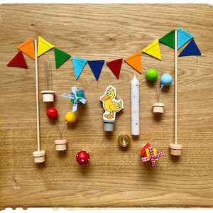 May include: A yellow duck cake topper with a rainbow flag bunting, a candle, and other cake decorations. The decorations include a red and yellow balloon, a red ball, a yellow ball, a red and white gift box, a red and white checkered box, and a red and white mushroom.