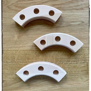 May include: Three light-coloured wooden curved blocks with evenly spaced holes. The blocks are arranged on a light brown wooden surface. The blocks have a smooth finish and a simple design. The blocks are likely for a building set or educational toy.