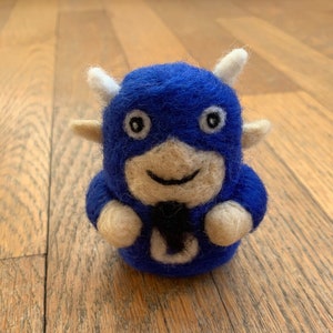 May include: A handmade felted blue bull figurine with white horns, ears, and facial details. The bull has a smiling face and a black tie-like shape on its chest. The toy is a whimsical, handcrafted item.