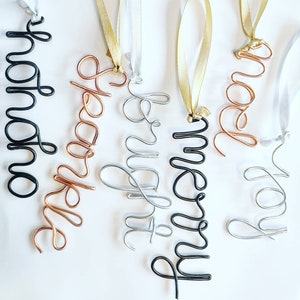May include: Five wire ornaments with different names written on them. The names are "Noah", "Sparkle", "Clement", "Merry", and "Joy". The ornaments are made of different coloured wire, including black, copper, silver, and gold.