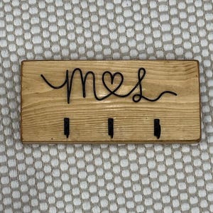 May include: A wooden key holder with the word "mrs" in black cursive script, with a heart replacing the "o". Three black hooks are below the text. The wood has a light brown finish.