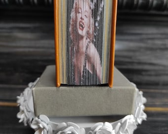 May include: A small, orange-brown book with a folded paper cover featuring a black and white image of Marilyn Monroe. The book is sitting on a gray box on a white pedestal with a floral design.
