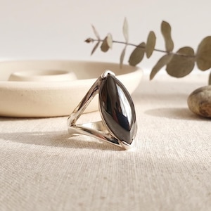 May include: A silver-toned ring featuring a large, smooth, marquise-shaped black stone set in a polished metal band. The band has a split design that tapers toward the base, securely holding the elongated, dark, reflective gemstone in place.