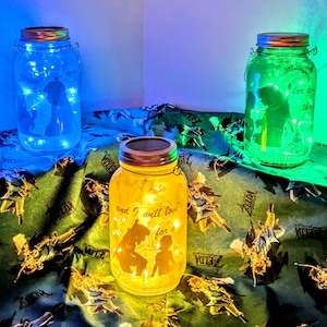Op de afbeelding: Drie glazen potten met lampjes erin, elk met een ander gekleurd licht. De potten zijn versierd met silhouetten en tekst. De tekst op de gele pot staat "and I will love you for a". De potten staan op een groene en zwarte stof met een herhalend patroon.