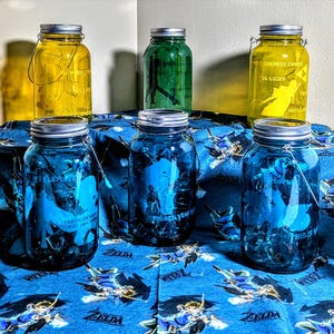 Op de afbeelding: Een verzameling van acht glazen potten met deksels, sommige zijn blauw en sommige zijn geel. De potten hebben verschillende ontwerpen, waaronder een personage uit een videogame. De potten staan op een blauw-witte gepatenteerde stof.