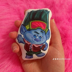 May include: A small, plush toy of a blue troll character with green hair, red goggles, and a red and brown outfit. The toy is held in a hand against a pink, fluffy background. The toy is a cartoon character.