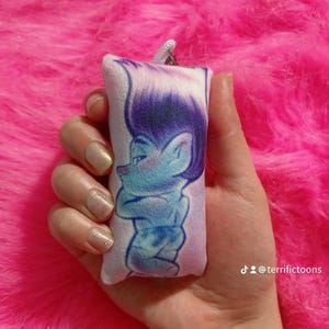 May include: A small, plush cushion featuring a purple and blue cartoon character. The character is a troll with blue skin, purple hair, and a blue and white outfit. The cushion is white with a light pink background. The cushion has a small loop at the top for hanging. The text "@terrifictoons" is visible in the bottom right corner of the image.