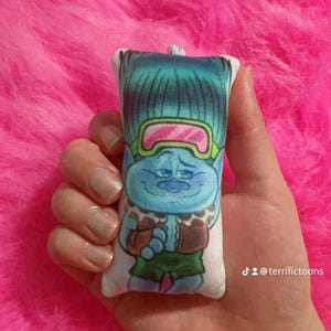 May include: A blue and green plush toy of a character from the Trolls movie wearing a brown vest, green trousers, and ski goggles. The toy is holding its hands in front of its chest. The toy is on a pink fluffy surface. The text "@terrifictoons" is visible in the bottom right corner of the image.