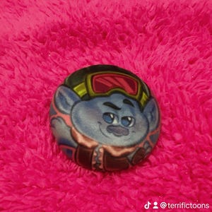 May include: A round, plush toy depicting a blue Troll character with goggles and a brown jacket. The toy is set against a bright pink, fuzzy background. The character has a smiling expression.