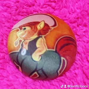 May include: A round button featuring a cartoon centaur character with red hair and a cowboy hat. The centaur is wearing a dark vest and is set against an orange and brown background. The button is on a bright pink, fuzzy surface.