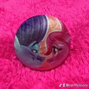 May include: A round, plush pillow featuring a cartoon illustration of two characters with blue and pink hair, touching noses. The pillow has a pink, fluffy background and the text "@terrifictoons" is visible.