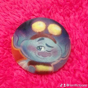 May include: A round, cartoon character button with a blue face, yellow eyes, and a brown mustache. The character is set against a vibrant pink background. The button has a winking expression.
