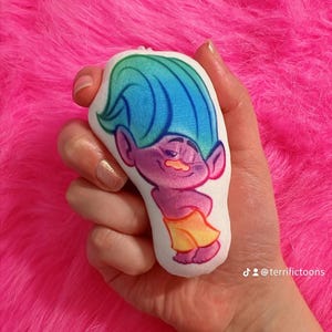 May include: A plush toy shaped like a troll with blue hair, a purple body, and a yellow skirt. The troll is smiling and has a moustache. The toy is held in a hand against a pink fluffy background. The text "@terrifictoons" is visible in the lower right corner of the image.