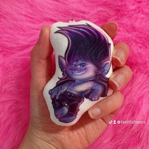 May include: A plush toy of a purple-haired troll with a blue body and black clothing. The troll is smiling and has a mischievous expression. The toy is held in a hand against a pink fluffy background. The text "@terrifictoons" is visible in the bottom right corner of the image.
