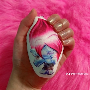 May include: A white, teardrop-shaped plush toy depicting a cartoon character with blue skin, pink hair, and brown shorts. The toy is held against a bright pink, fluffy background. The character has a small earring and a belt buckle. The Instagram handle @terrifictoons is visible.