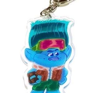 May include: A blue troll character with green hair wearing a brown jacket, green pants, and goggles. The character is holding a brown object in their right hand. The character is on a clear acrylic keychain.