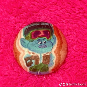 May include: A circular badge with a cartoon character in blue, wearing goggles and a green outfit. The badge has a brown and orange striped border, set against a bright pink, fluffy background. The text "@terrifictoons" is at the bottom.