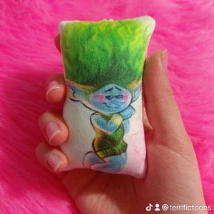 May include: A small, white plush pillow featuring a printed green-haired blue troll character. The troll is wearing a green shirt and has a grumpy expression.