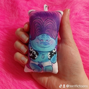 May include: A purple and blue plush keychain featuring a cartoon troll with a purple mohawk, wearing a white shirt with a black and white floral pattern. The keychain is held in a hand.