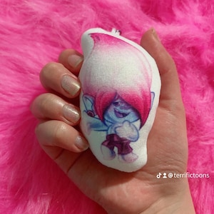 May include: A plush toy of a pink-haired troll with blue skin and a white shirt. The troll is holding a heart in its hands. The toy is on a pink fuzzy surface.  The text "@terrifictoons" is visible in the bottom right corner of the image.