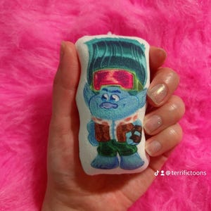 May include: A blue and green plush toy with a white background. The toy features a character with blue skin, green hair, and a red and green headband. The character is wearing a brown jacket and green trousers. The toy is holding a brown ball. The text "@terrifictoons" is visible in the bottom right corner of the image.