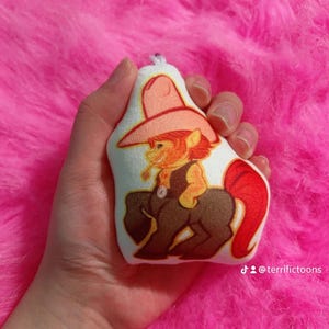 May include: A plush toy shaped like a horse wearing a cowboy hat and waistcoat. The horse is brown and white with a red tail. The toy is holding a cigarette in its mouth. The toy is on a pink fluffy surface. The text "@terrifictoons" is visible in the bottom right corner of the image.