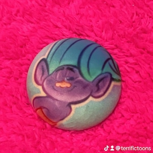 Pode incluir: Um design de personagem de desenho animado redondo, roxo e verde-azulado, sobre um fundo azul claro. O personagem tem orelhas grandes e um rosto sorridente. O design está sobre um fundo rosa brilhante e difuso. O texto "@terrifictoons" é visível.