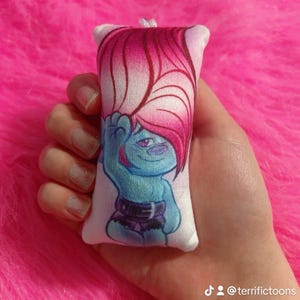 May include: A small, white cushion with a blue and pink cartoon character printed on it. The character has pink hair and is wearing a black belt and shorts. The cushion is being held in a hand.