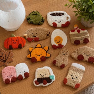 May include: A collection of small, tufted punch needle coasters featuring various cute characters and food items, including a pumpkin, a sun, a frog, a croissant, a fried egg, a coffee mug, a peanut, a leaf, and other whimsical shapes with smiling faces.