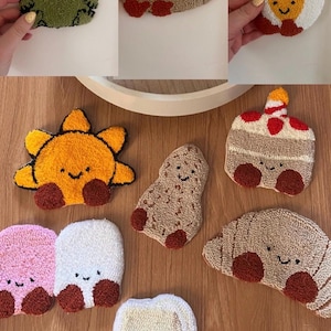 May include: A selection of handmade, tufted coasters in various shapes and colours. Designs include a green frog, a brown mole, a fried egg, a smiling sun, a peanut, a slice of cake, a croissant, and other cute characters. Each coaster has a smiling face.