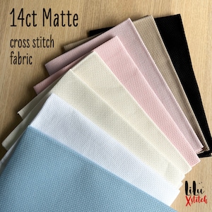 May include: A variety of 14 count matte Aida cross stitch fabric in different colors, including black, beige, light pink, white, cream, and light blue. The fabric is folded and stacked on a wooden surface. The text "14ct Matte cross stitch fabric" is visible at the top of the image.