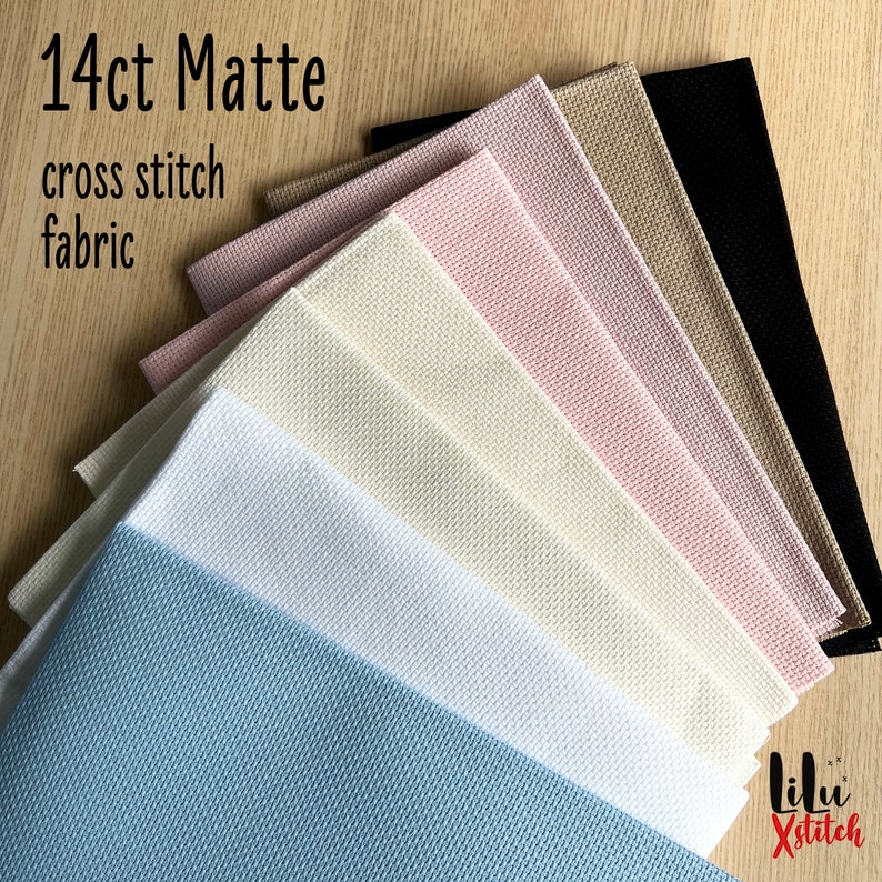 May include: A variety of 14 count matte Aida cross stitch fabric in different colors, including black, beige, light pink, white, cream, and light blue. The fabric is folded and stacked on a wooden surface. The text "14ct Matte cross stitch fabric" is visible at the top of the image.