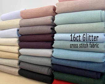 Può includere: Una pila di tessuto per punto croce glitter da 16 fili, in vari colori, tra cui rosa, beige, blu, grigio, verde e rosso. Il tessuto è una trama testurizzata con un leggero luccichio. Il testo "16ct Glitter cross stitch fabric" è stampato sul tessuto.