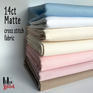 May include: A stack of 14 count matte cross stitch fabric in various colors, including light blue, white, cream, beige, and pink. The fabric is folded and stacked neatly. The text "14ct Matte cross stitch fabric" is visible on the left side of the image.