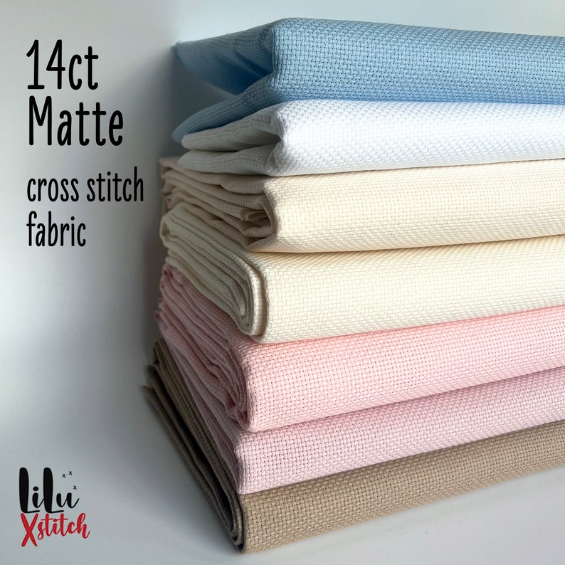 May include: A stack of 14 count matte cross stitch fabric in various colors, including light blue, white, cream, beige, and pink. The fabric is folded and stacked neatly. The text "14ct Matte cross stitch fabric" is visible on the left side of the image.