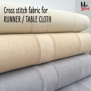 May include: Three rolls of cross stitch fabric in cream, beige, and pale grey. The fabric is labelled "Cross stitch fabric for RUNNER / TABLE CLOTH". The fabric has a striped design with fringed edges, suitable for crafting.