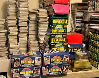 May include: A large collection of vintage baseball cards, including unopened packs and boxes, stacked on a table. The cards are from the 1991 Score series and feature various players and teams. There are also several boxes of Ultra Baseball cards and a gold-colored trophy.